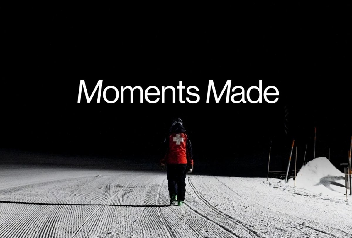 A person in a red jacket with a white cross walks alone on a snow-covered path at night; text above reads "Moments Made.