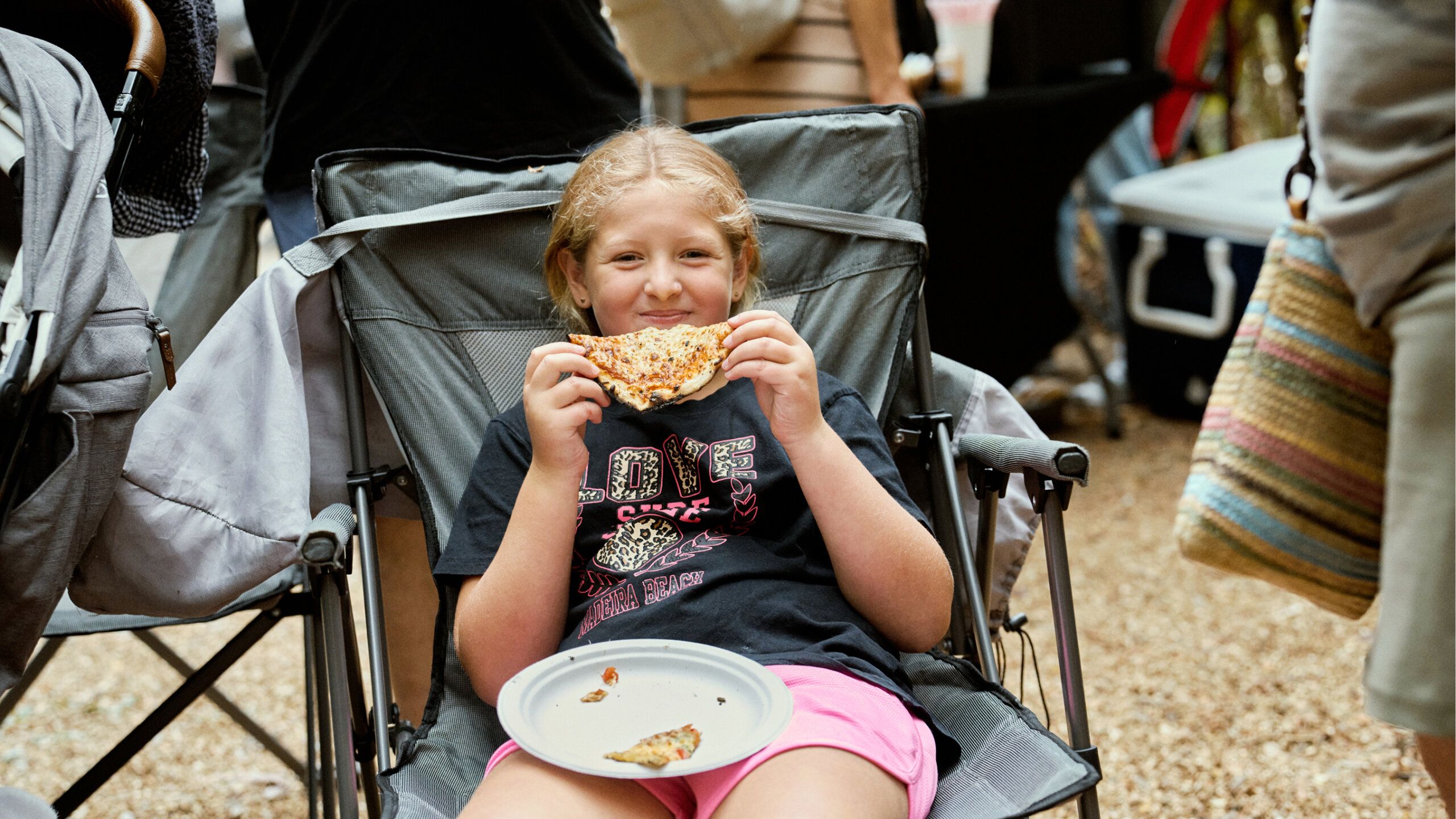A young girl sits in a camping chair outdoors, holding a slice of pizza and a paper plate with pizza crusts, smiling at the camera.