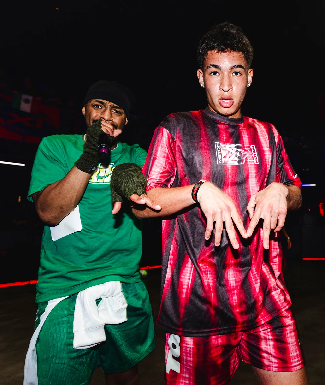 Two young men on stage; one in a green outfit holding a mic, the other in a red sports jersey making a hand gesture. Both have expressive faces and are under stage lighting.