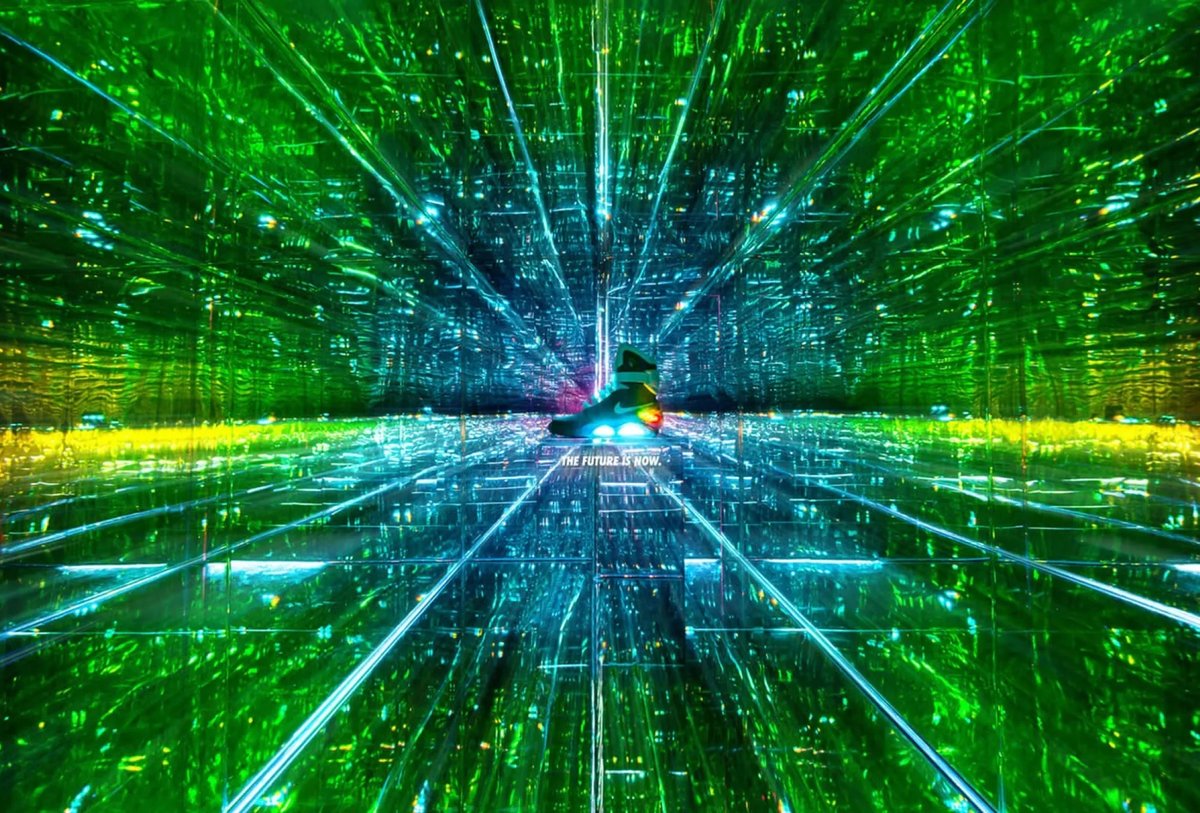 A person stands in the center of a mirrored room filled with green and blue lights, creating an illusion of infinite reflections and depth.