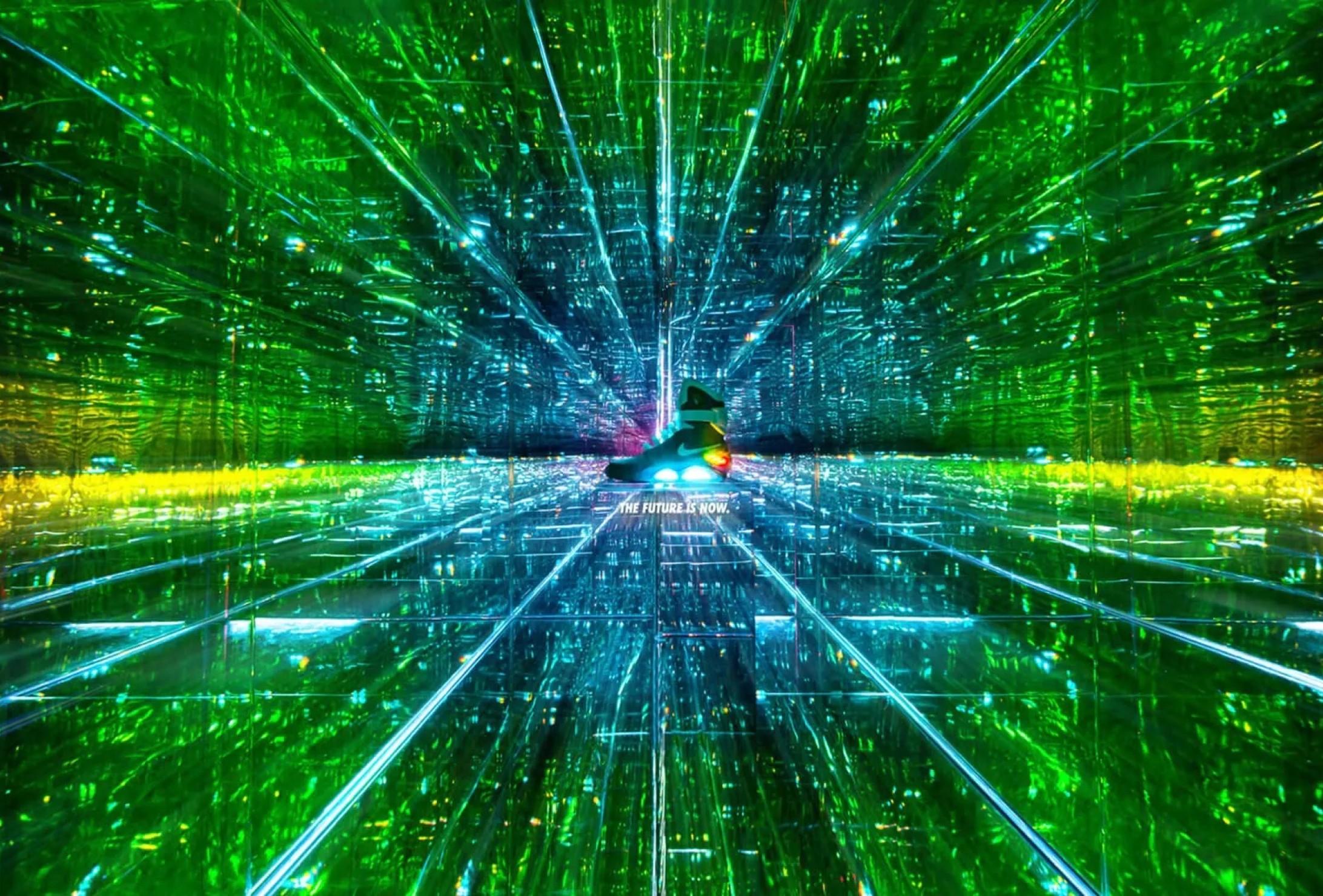 A person stands in the center of a mirrored room filled with green and blue lights, creating an illusion of infinite reflections and depth.