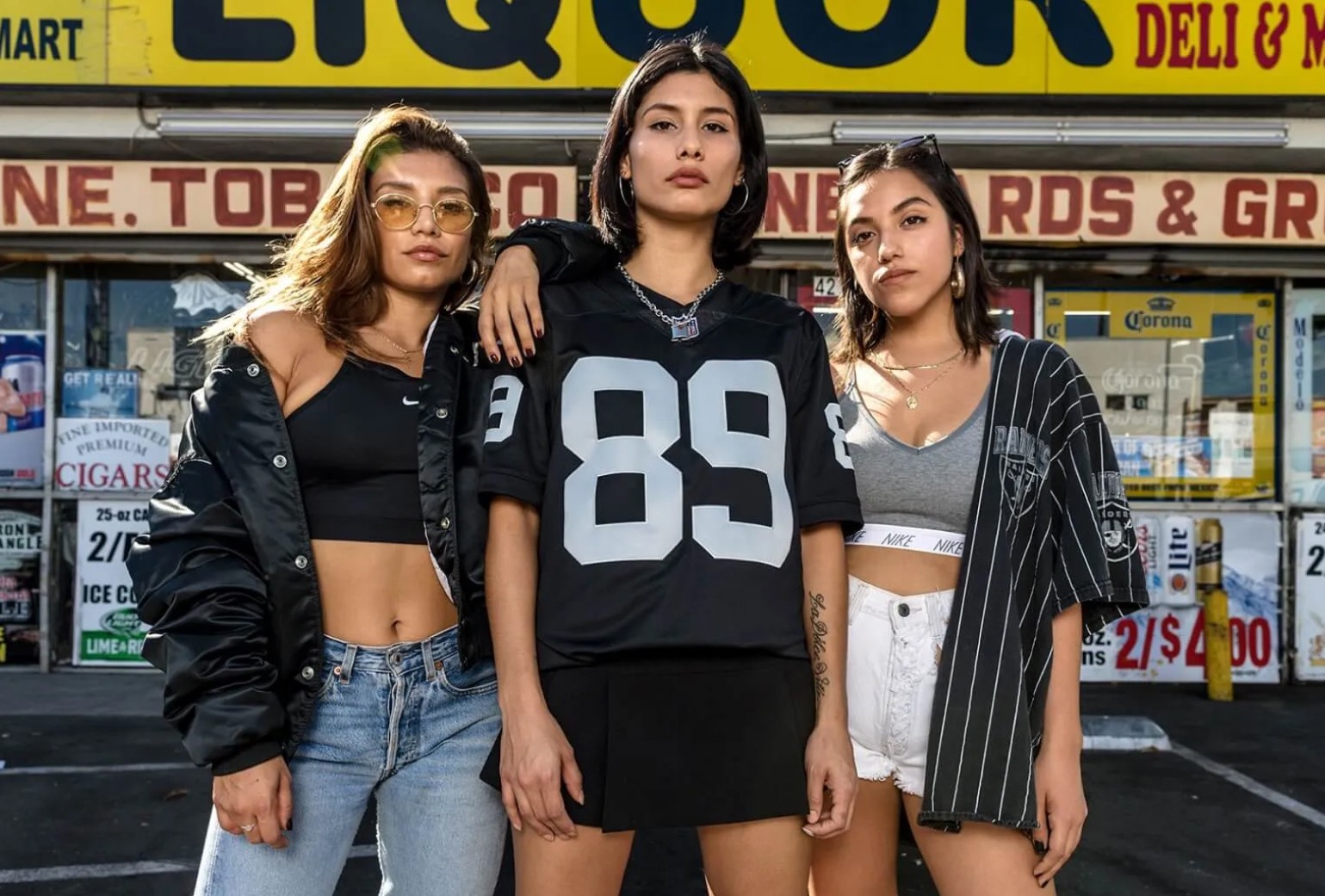 Three young women pose confidently in front of a liquor store, wearing casual, sporty outfits with a yellow store sign visible in the background.