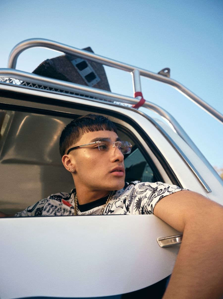 A person with short hair and glasses leans out of a car window, looking to the side. The car has a roof rack and the sky is clear.