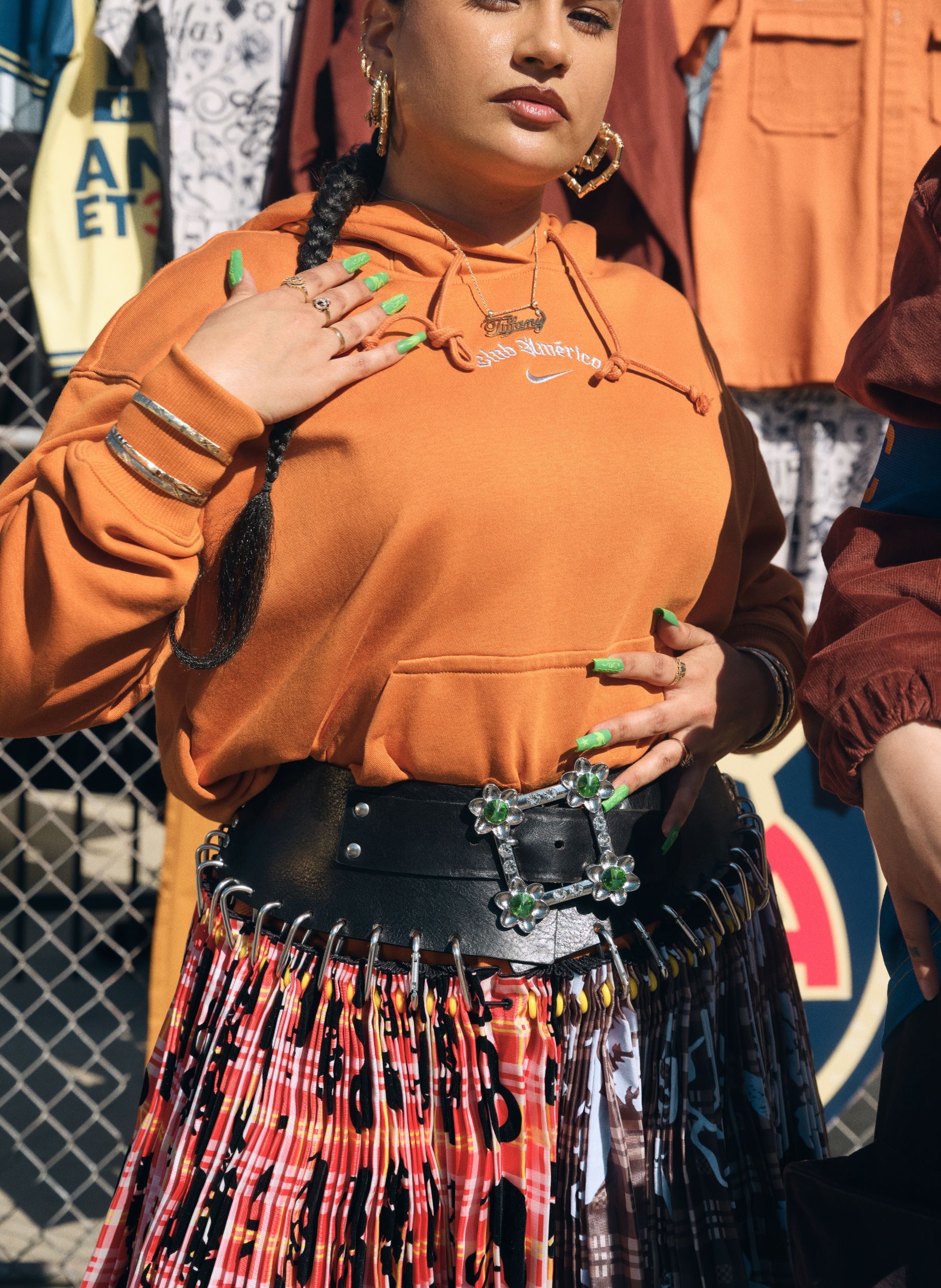 A person wearing a burnt orange hoodie, patterned skirt, chunky belt with jeweled buckle, gold jewelry, and green nail polish stands outdoors with one hand on their chest.