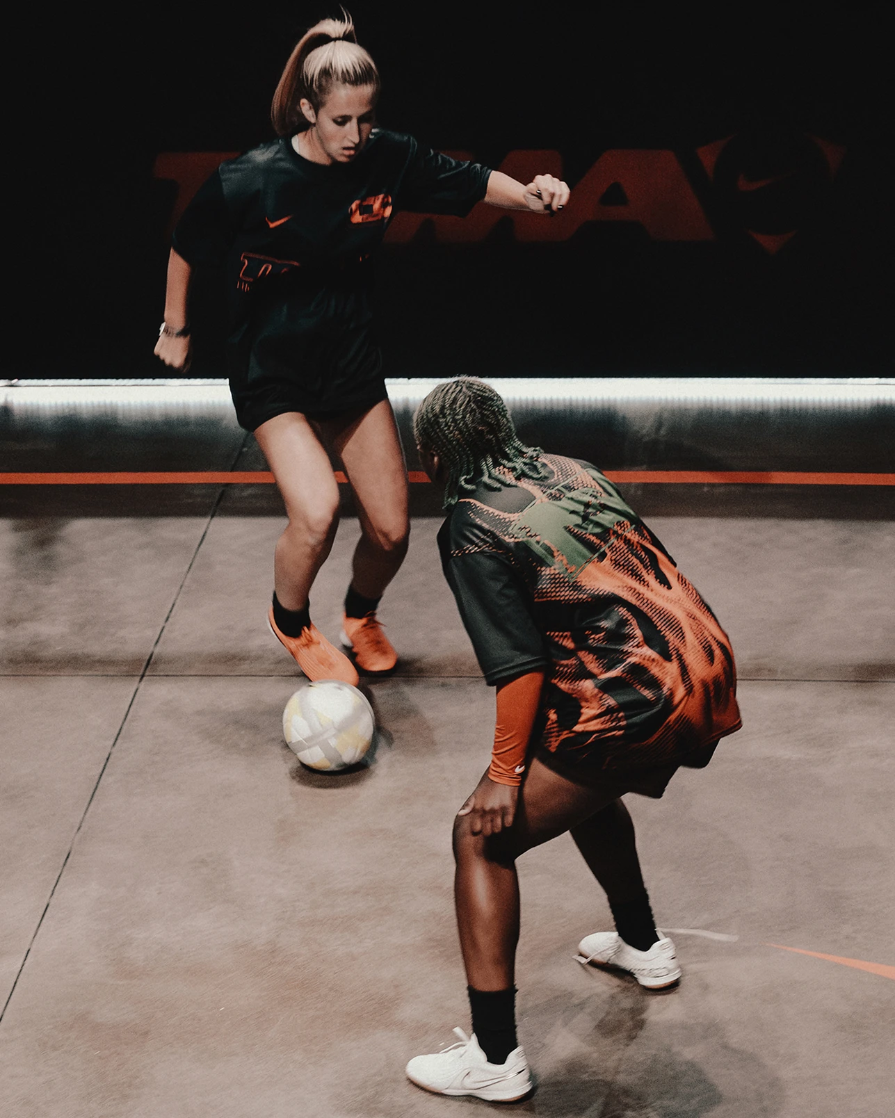 Two women in athletic clothing play soccer indoors, with one dribbling the ball while the other defends.