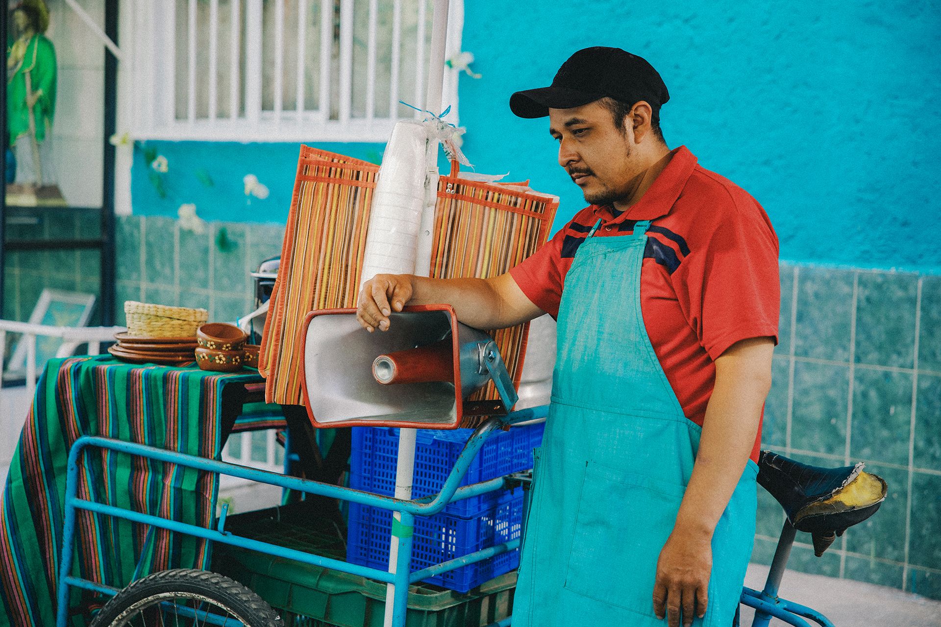 A man wearing a red shirt, turquoise apron, and black cap stands by a street vendor cart equipped with various items including a red megaphone, baskets, and a blue crate in an urban setting.