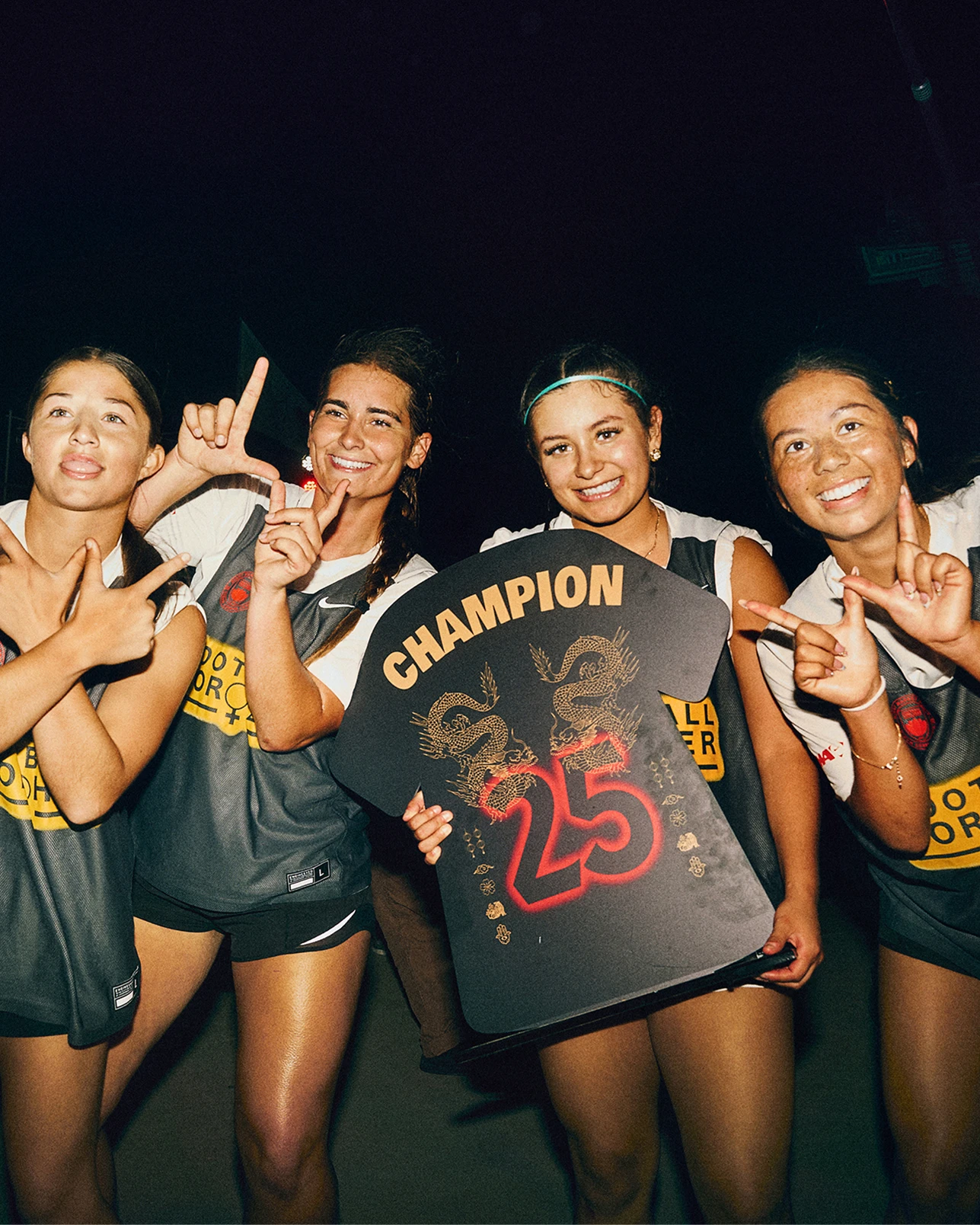 Four women in sports uniforms pose together, one holding a black sign with “CHAMPION 25” and dragon designs. They are smiling and making hand gestures, standing against a dark background.
