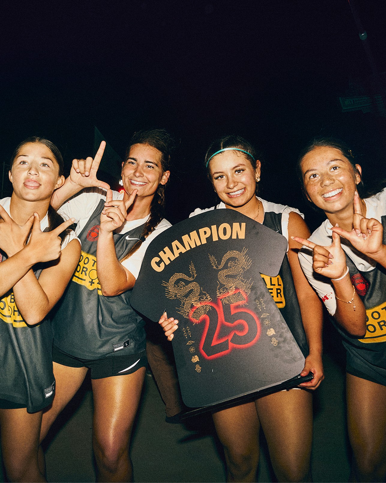 Four women in sports uniforms pose together, one holding a black sign with “CHAMPION 25” and dragon designs. They are smiling and making hand gestures, standing against a dark background.