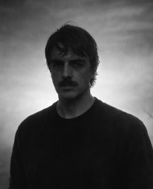 A man with a mustache and dark hair stands outdoors at dusk or dawn, with a bare tree and cloudy sky in the background.