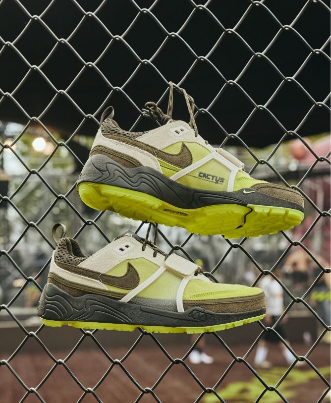 A pair of green and brown Nike sneakers with yellow soles is hanging by the laces on a chain-link fence, with a blurred outdoor background.