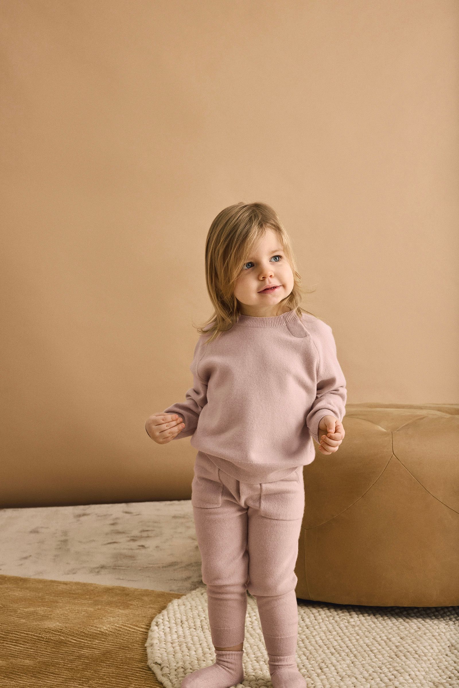 A young child wearing a light pink sweater and pants stands on a patterned rug with a tan pouf and beige wall in the background.