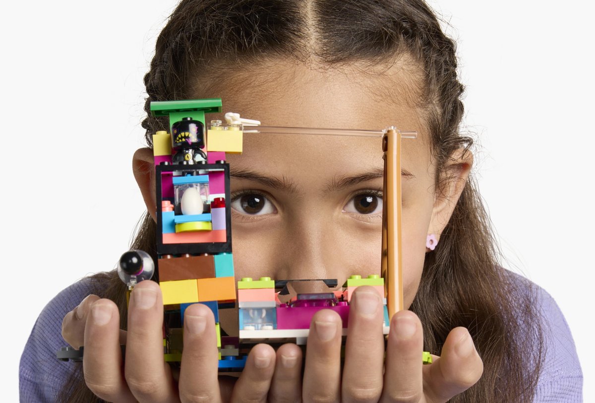 A girl holds a colorful LEGO structure in both hands, with her face partially visible behind it against a white background.