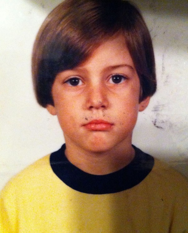 A young child with straight brown hair and a neutral expression wears a yellow shirt with dark trim, facing the camera against a plain background.