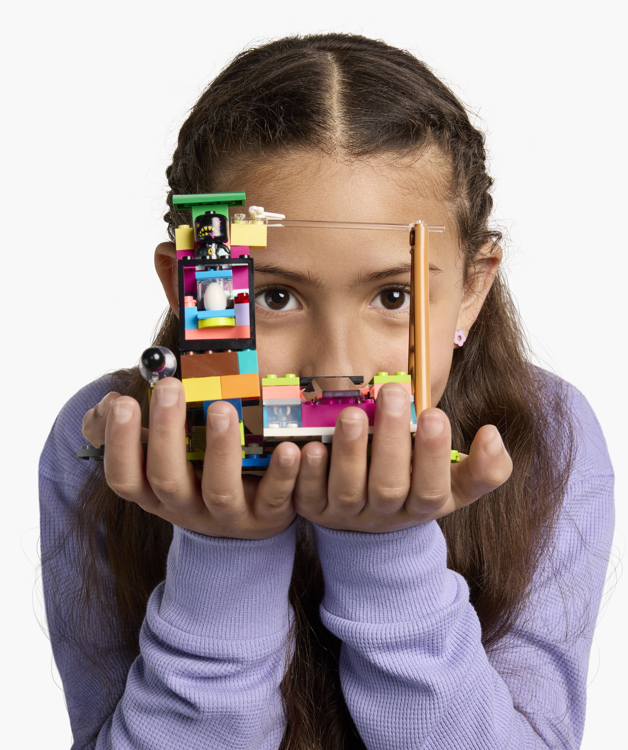 A young girl wearing a purple sweater holds a colorful LEGO creation up to her face with both hands, while staring directly at the camera.