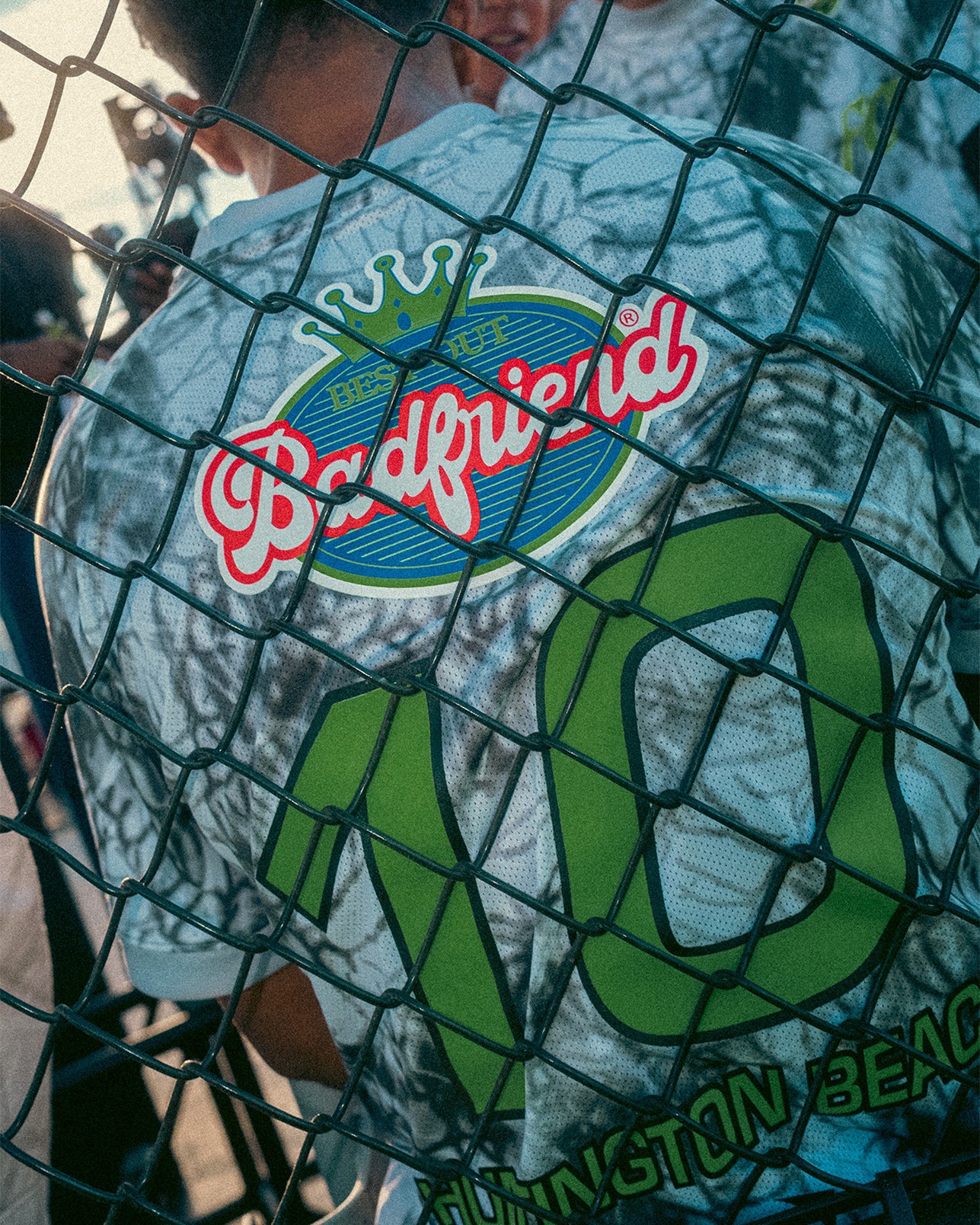 Person wearing a tie-dye shirt that says “Badfriend” and “10 Huntington Beach” stands behind a green chain-link fence.