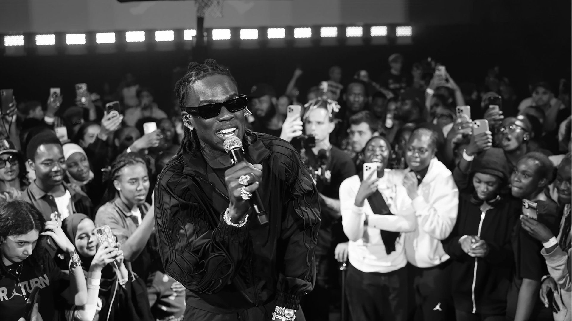 A performer in sunglasses sings into a microphone on stage, surrounded by an enthusiastic crowd taking photos and videos with their phones.