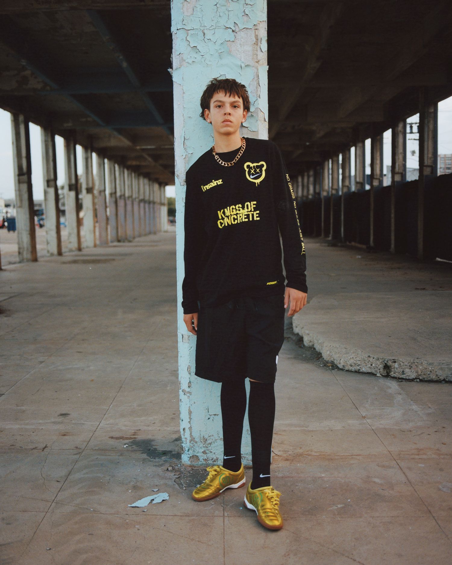 A young person stands on a concrete floor under a partially open, worn-down structure, wearing black athletic gear and gold soccer shoes.