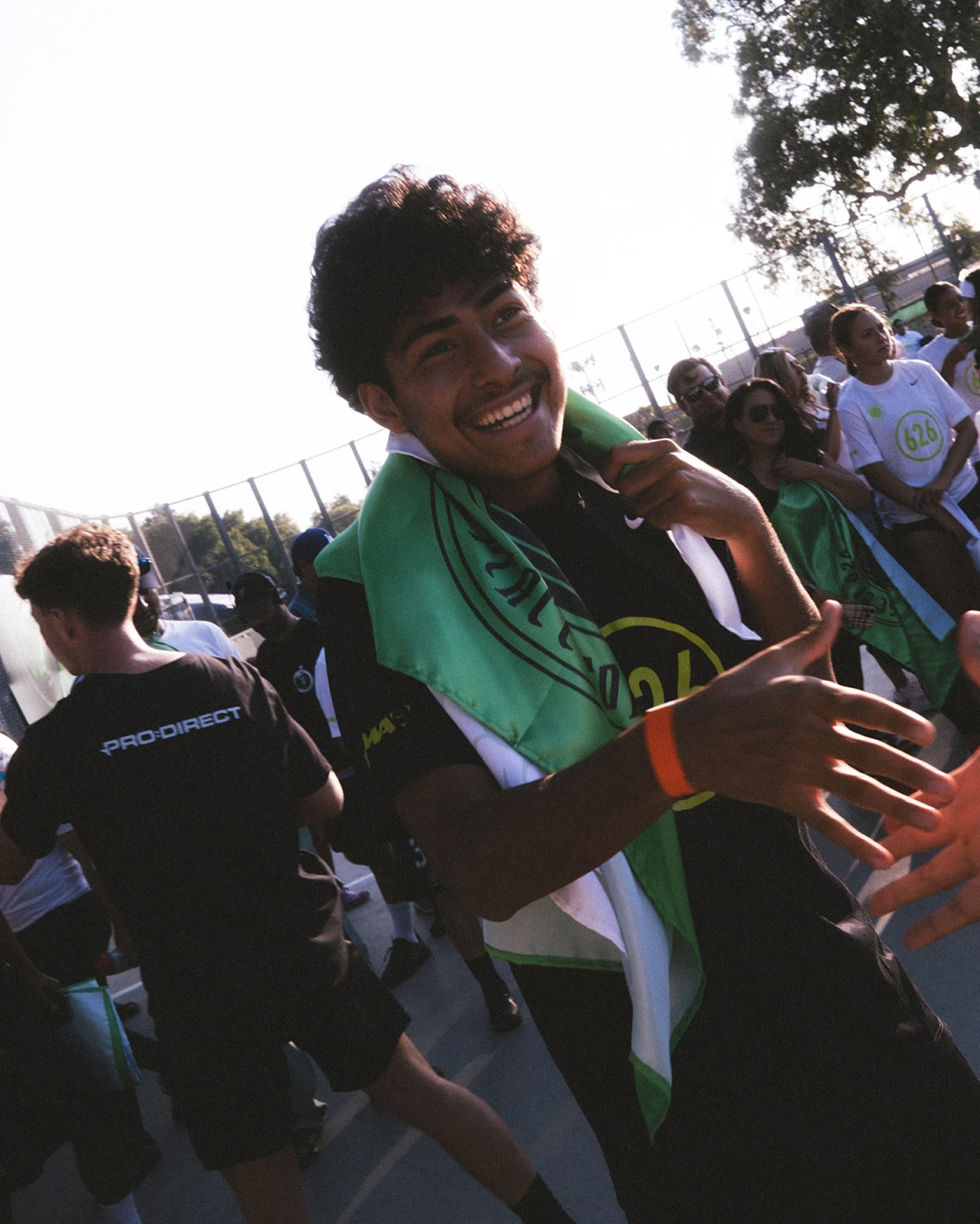 Smiling person draped in a green and white cloth extends a hand while standing on an outdoor court with a group of people in the background.