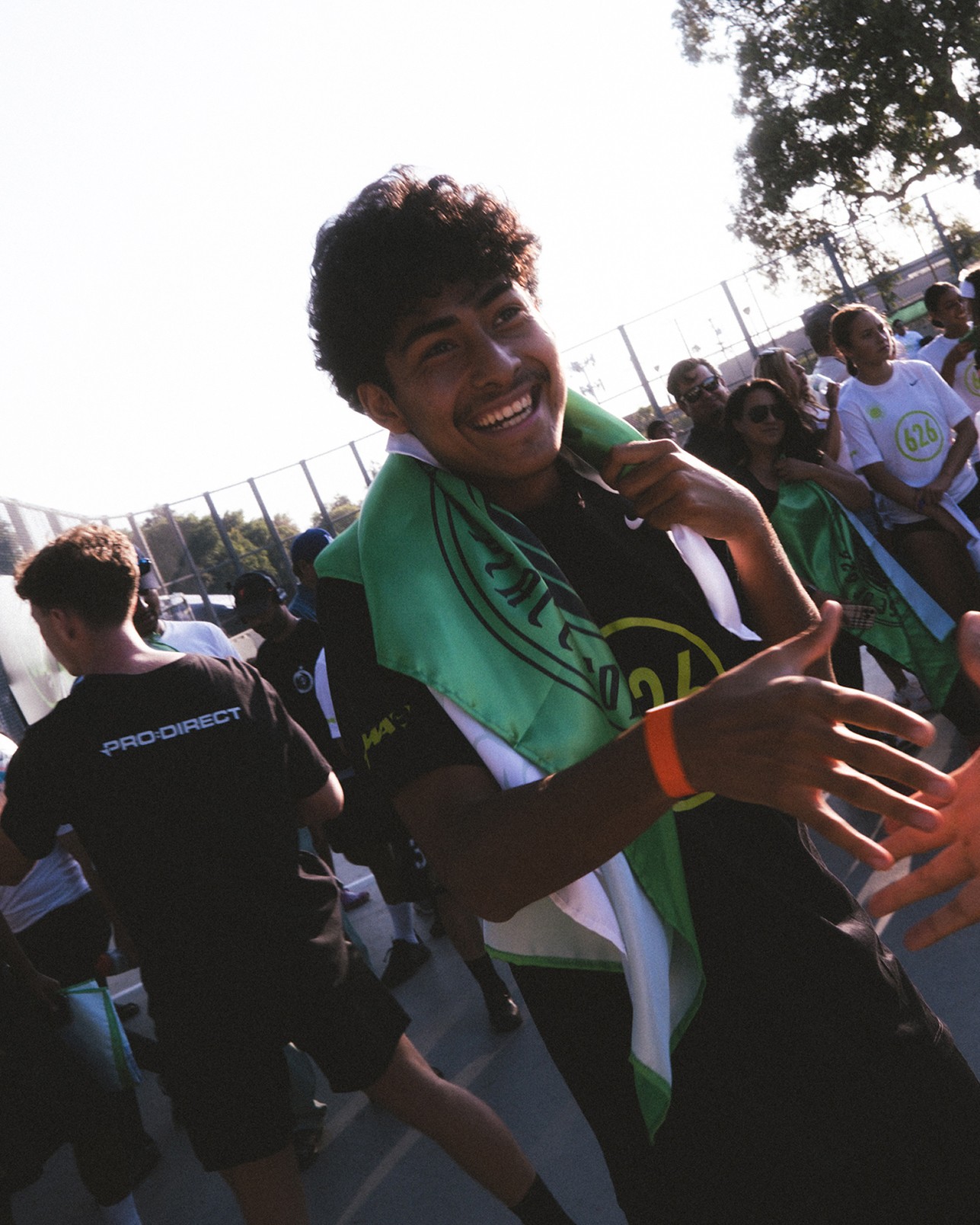 Smiling person draped in a green and white cloth extends a hand while standing on an outdoor court with a group of people in the background.