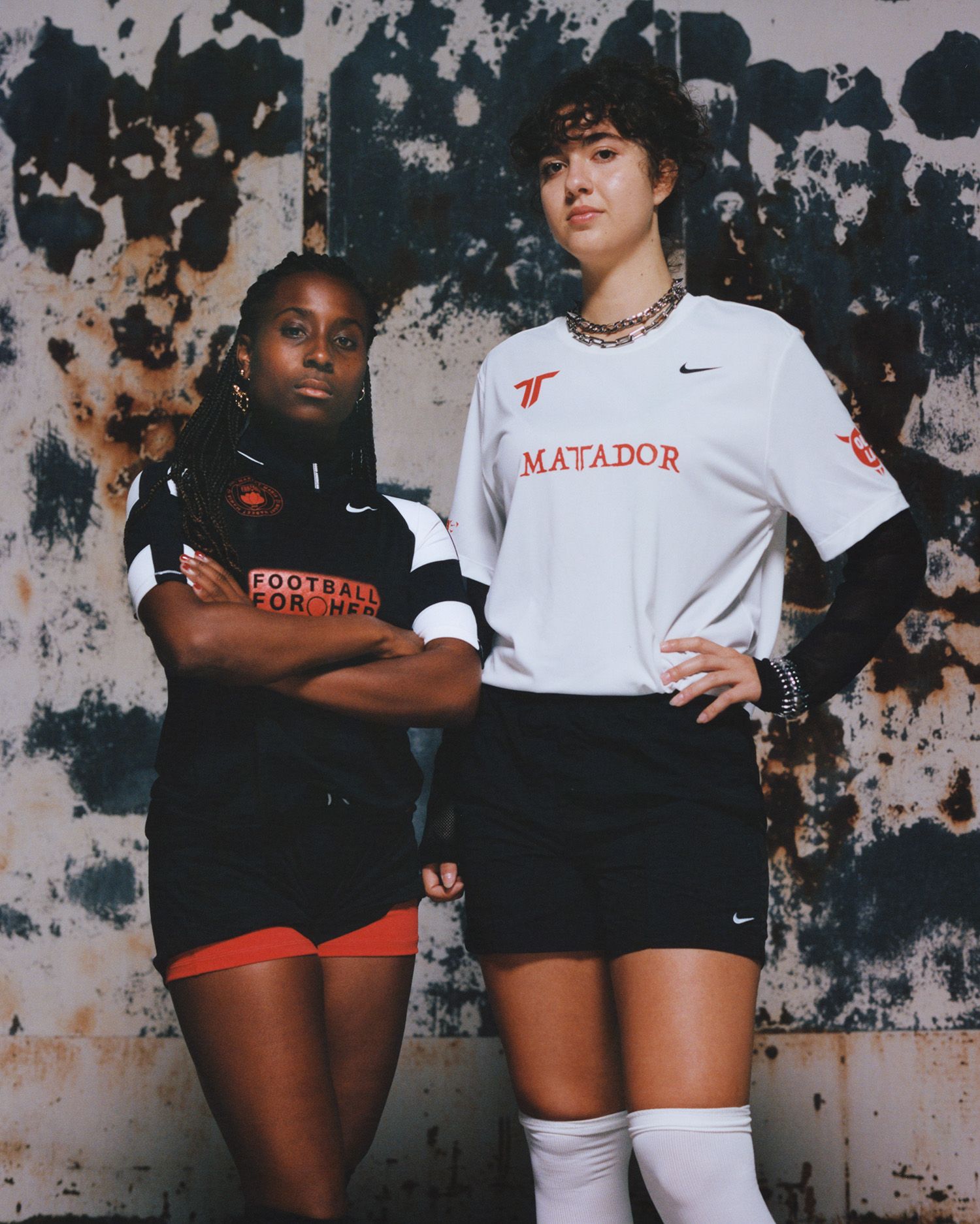 Two women in soccer uniforms stand side by side against a weathered, textured wall, looking confidently at the camera.