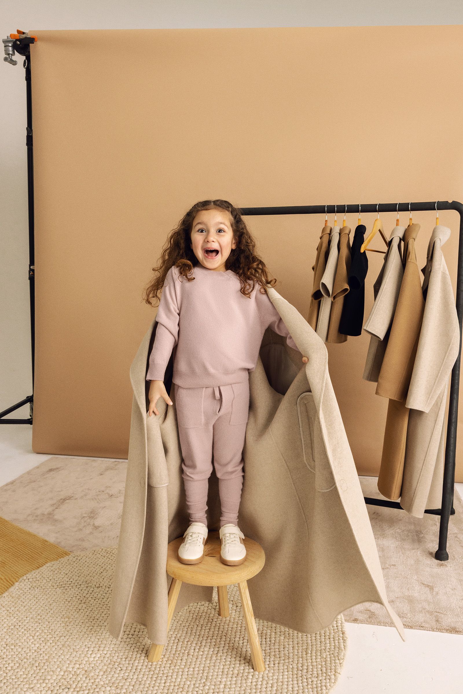 A young child stands on a wooden stool, draped in a beige coat, with neutral-toned clothing hanging on a rack behind them.