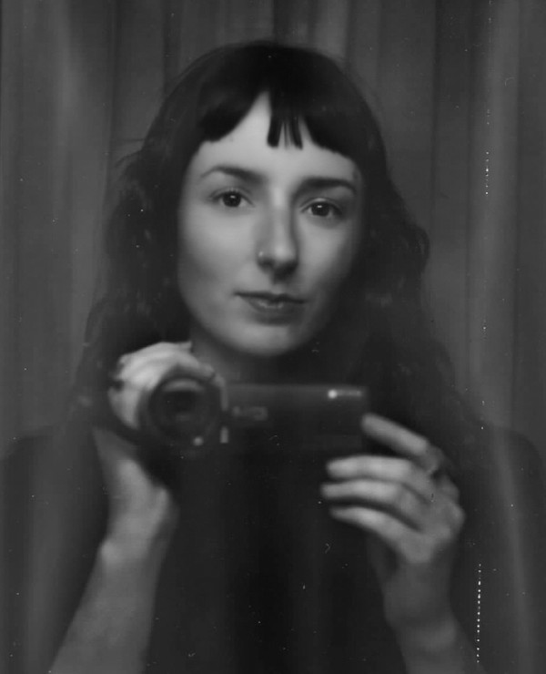 A person with dark hair and bangs holds a camera, facing a mirror. The photo is in black and white with a soft, slightly blurred effect.