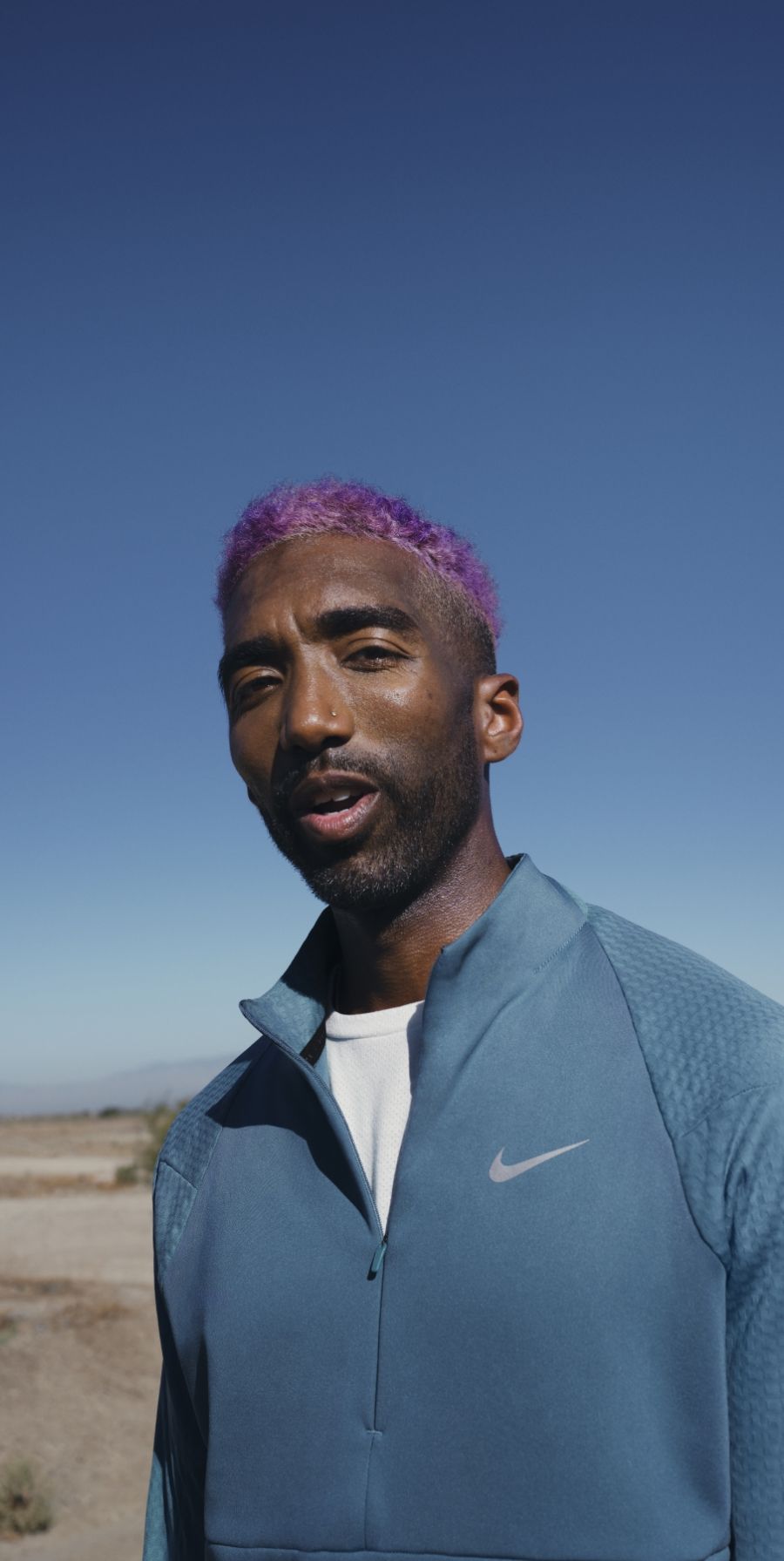 Runner with purple hair and blue running jacket looking into camera with desert and blue sky behind him