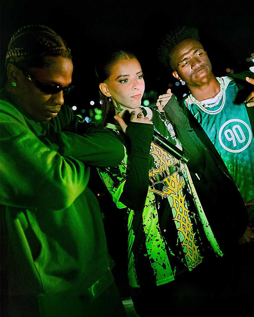 Three young adults pose at night under green lighting, wearing casual streetwear including hoodies, a patterned shirt, and a sports jersey, with a cityscape blurred in the background.