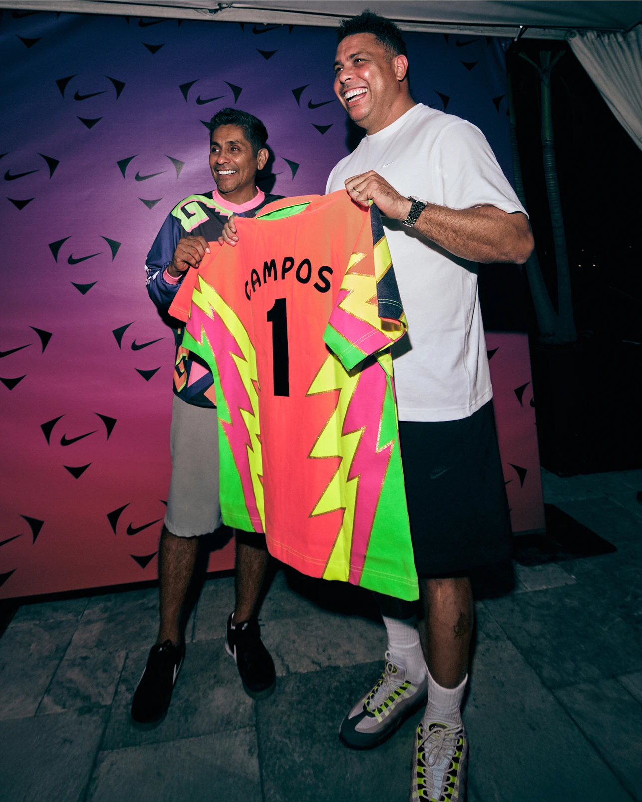 Two men stand smiling, holding a bright neon soccer jersey with “CAMPOS 1” on the back, in front of a purple patterned backdrop.