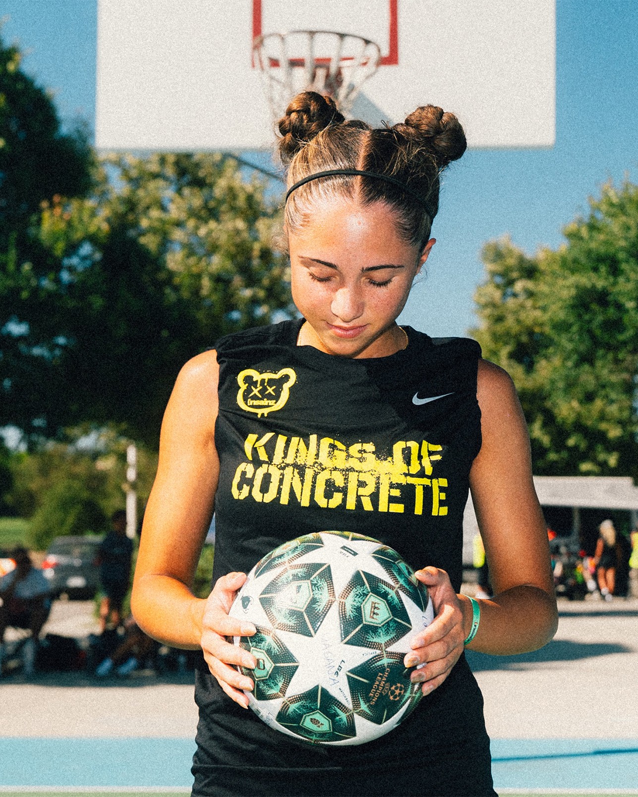 A young woman with braided buns holds a soccer ball and looks down, standing on an outdoor basketball court. She wears a “Kings of Concrete” sleeveless shirt. Trees and people are in the background.