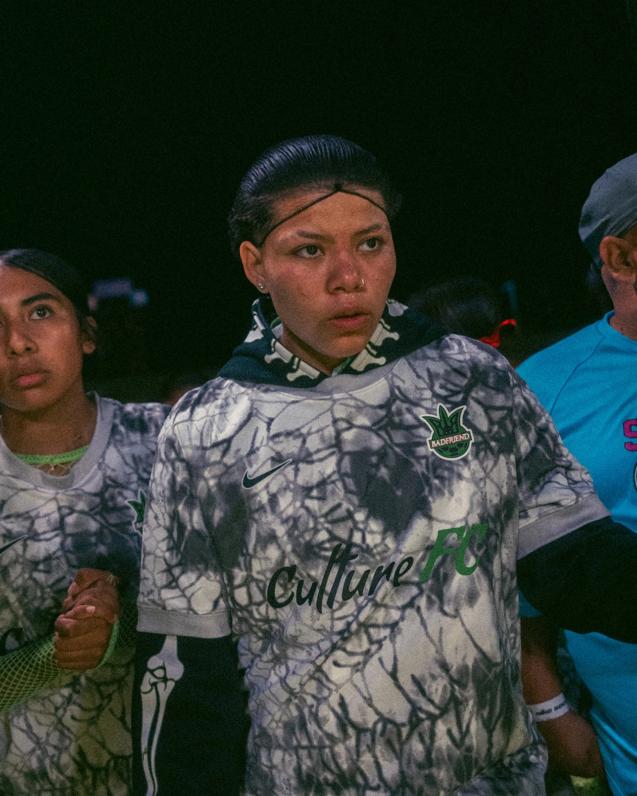 Two young people in marbled gray soccer jerseys stand closely together at night, one holding hands with someone off-frame, both looking ahead with serious expressions.