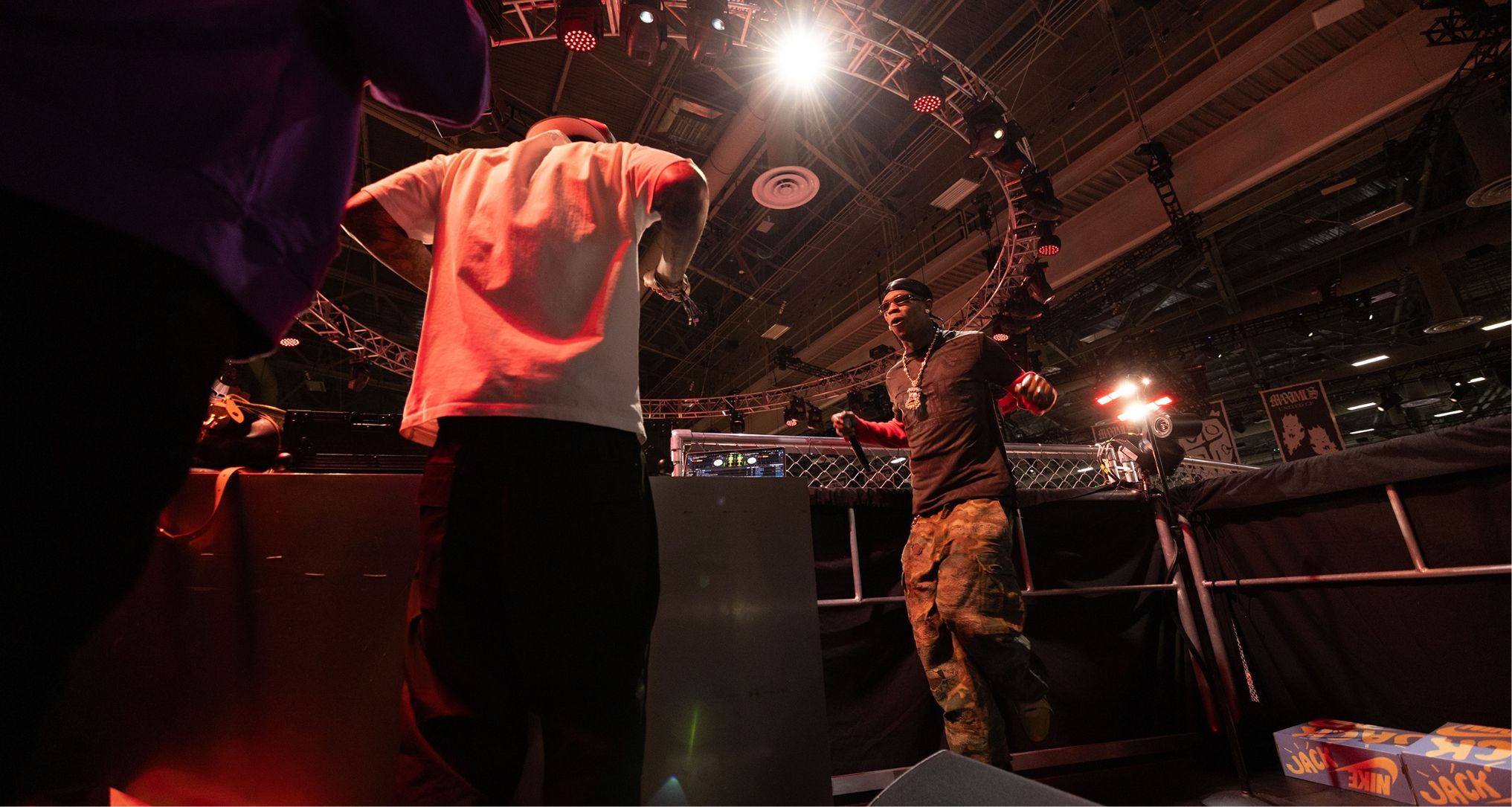 Two people including Travis Scott are on stage near a DJ setup with a backdrop of lights and equipment in a performance setting.