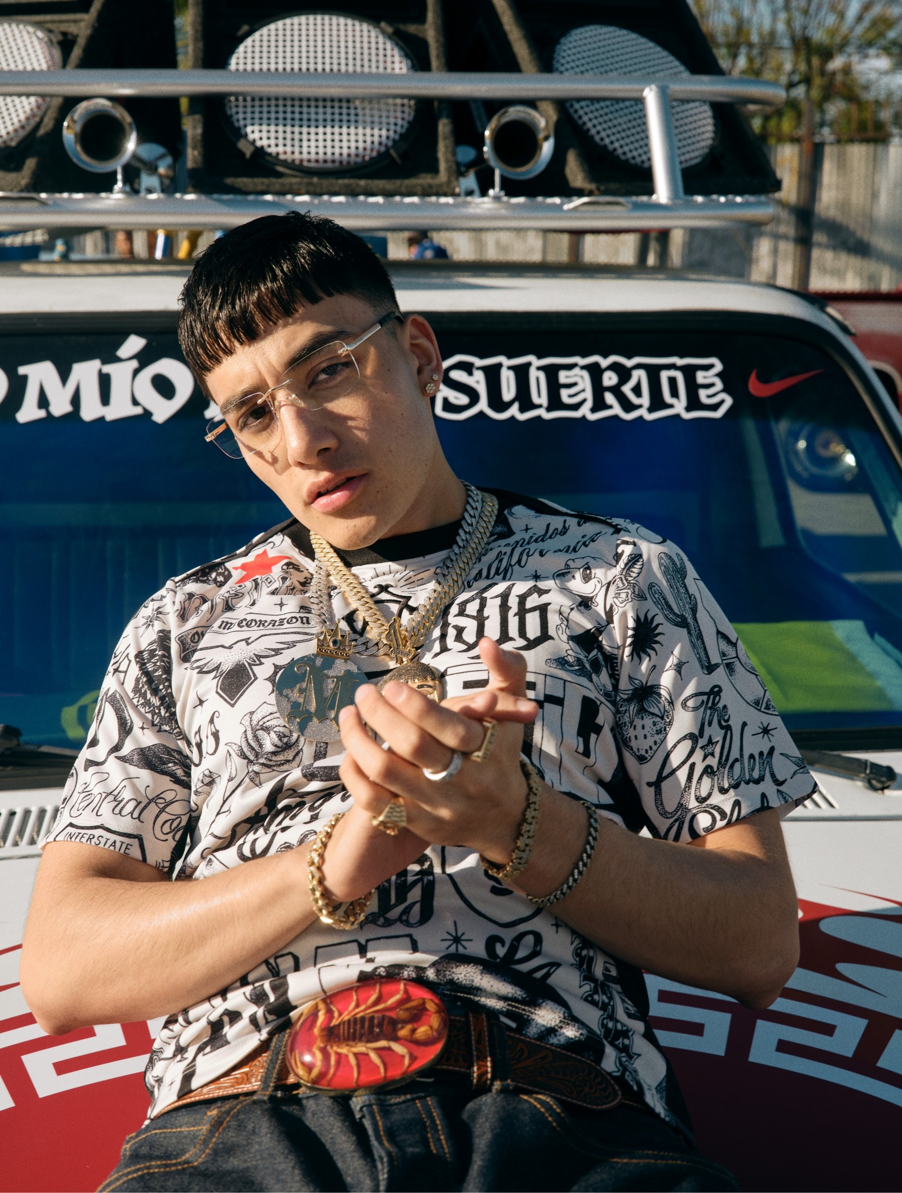 A young man wearing glasses, a graphic t-shirt, and gold jewelry poses in front of a decorated car with large speakers on the roof.