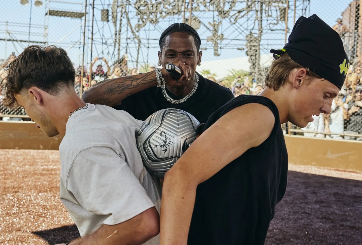 A man holds a microphone while two people balance a soccer ball between their backs on a dirt field, with a metal structure and crowd in the background.