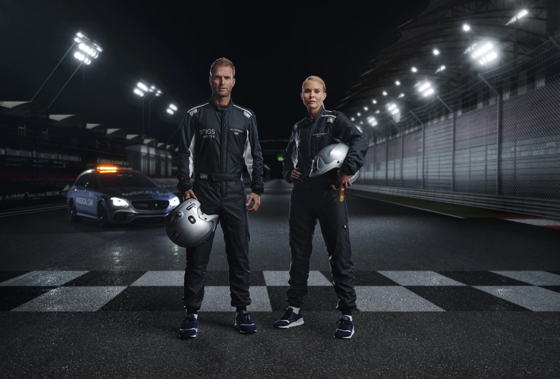 Two race car drivers in black racing suits stand on a wet racetrack at night, holding helmets, with a safety car and lit grandstands in the background.