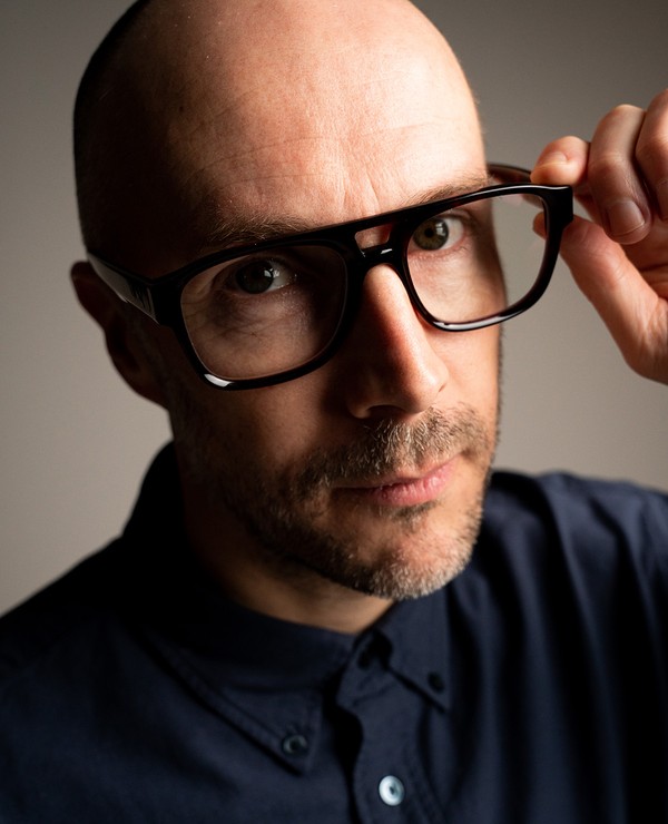 A man with a shaved head and stubble adjusts his large black glasses while looking at the camera. He is wearing a dark button-up shirt.