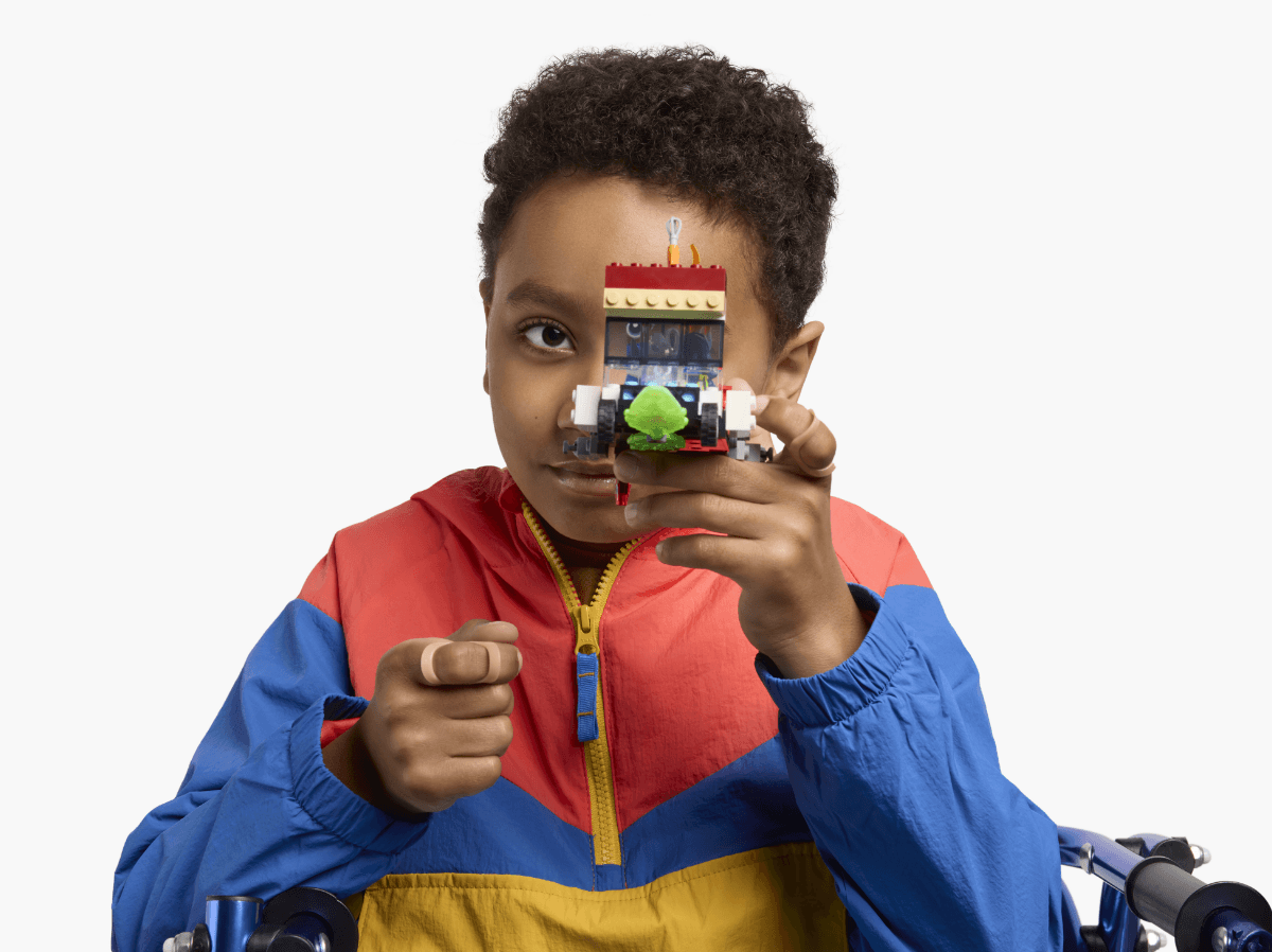 A child in a colorful jacket holds up a LEGO creation with one hand, covering part of their face while smiling.