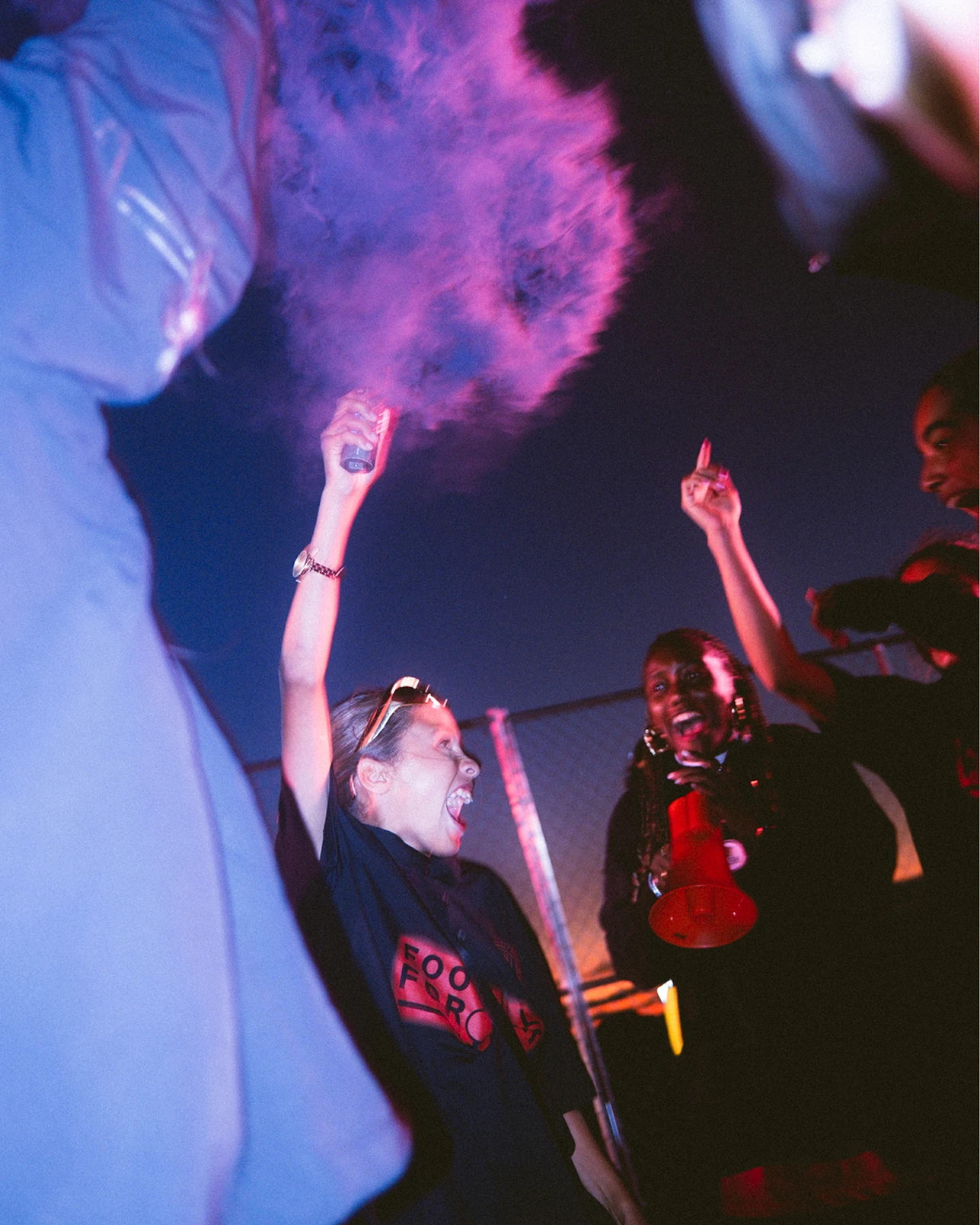 People gather outdoors at dusk, cheering and holding a smoke bomb emitting purple smoke. One person holds a megaphone. The mood appears energetic, with a fence and evening sky in the background.