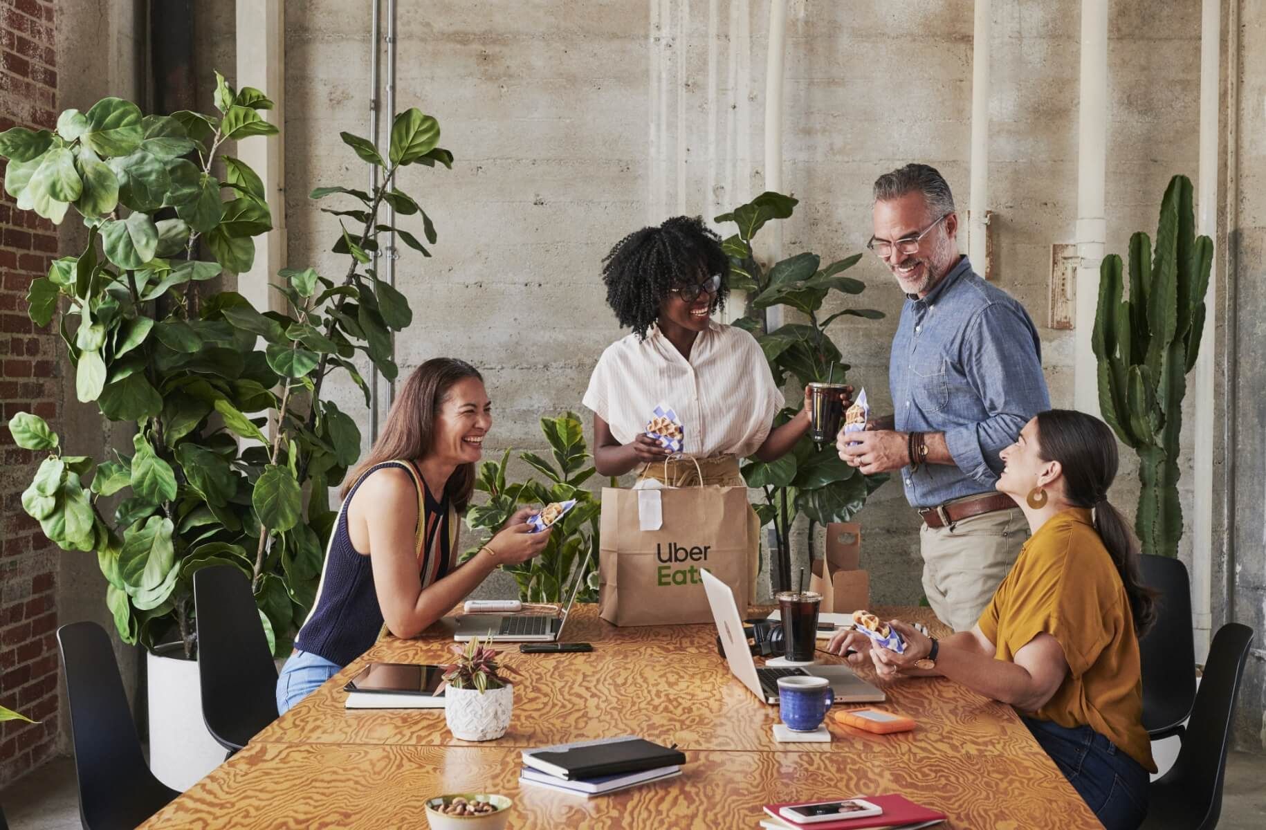 office meeting with laptops and note books on table along with take out waffles from Uber eats. Four people sitting and standing around table are laughing and smiling