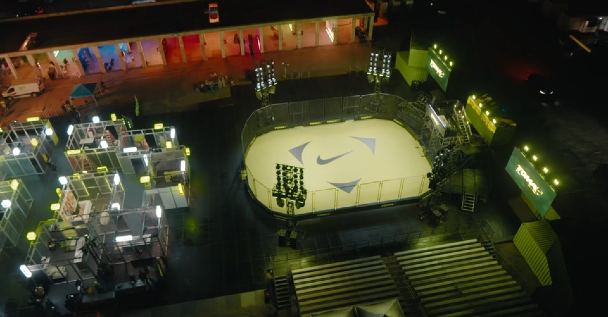 A nighttime aerial view of an illuminated outdoor sports court with a Nike logo at the center, surrounded by empty bleachers and event lighting.
