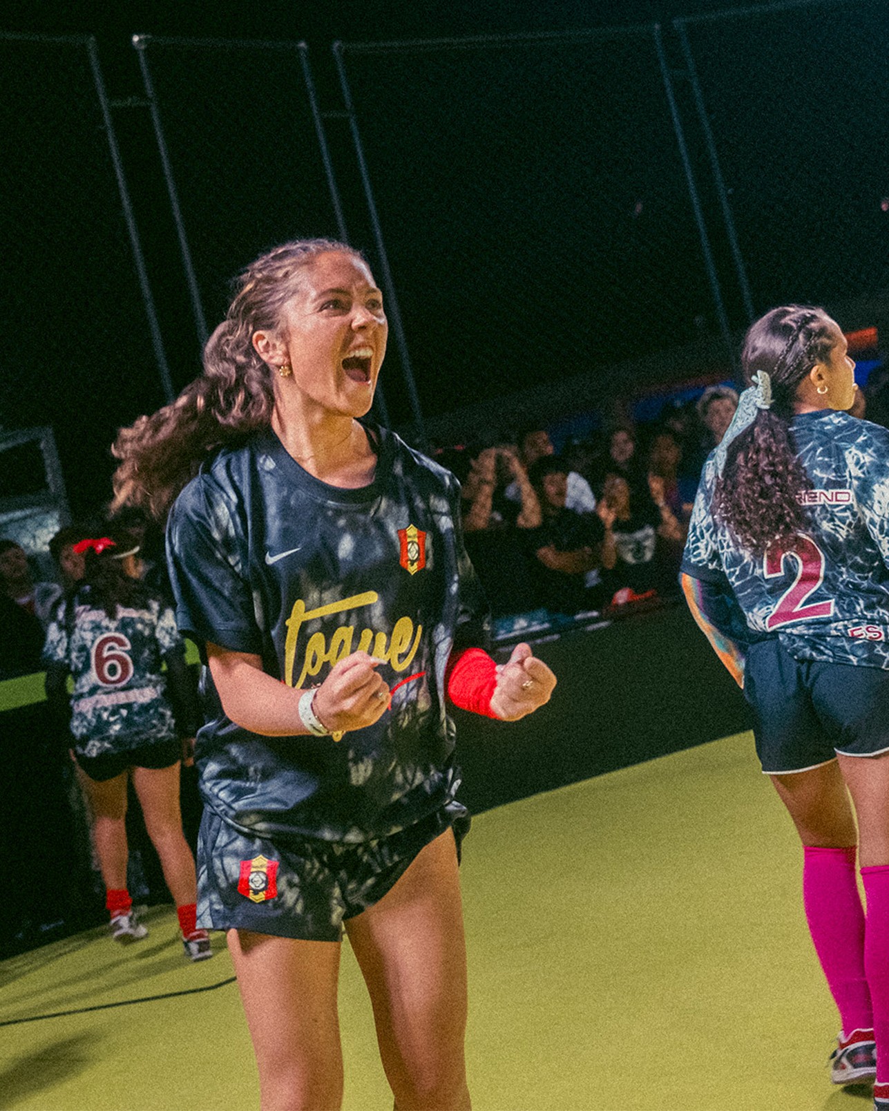A young woman in a black soccer uniform clenches her fists and shouts in celebration on an indoor field, as teammates and spectators look on.