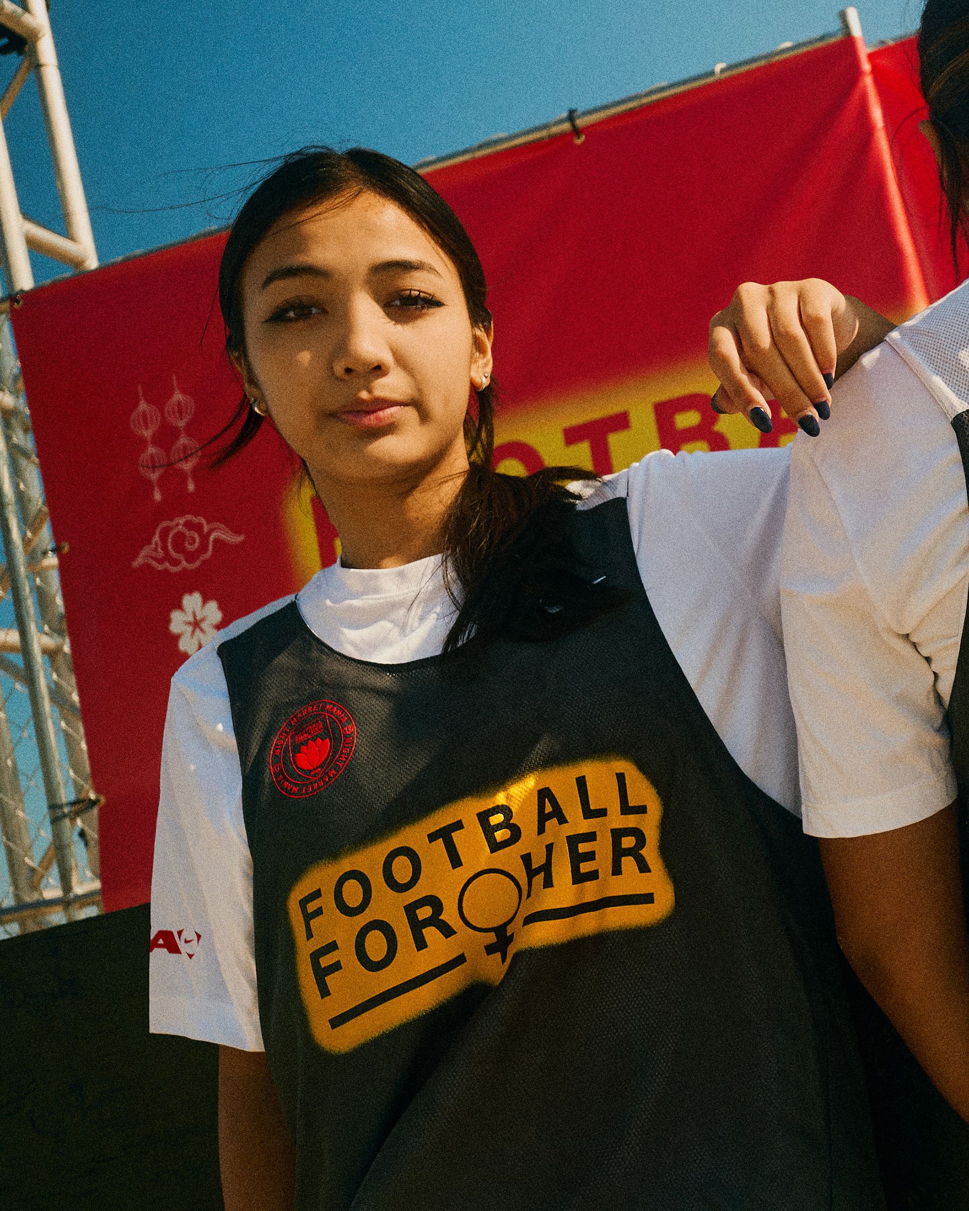 A young woman wearing a "Football For Her" vest stands in front of a red banner, looking at the camera.