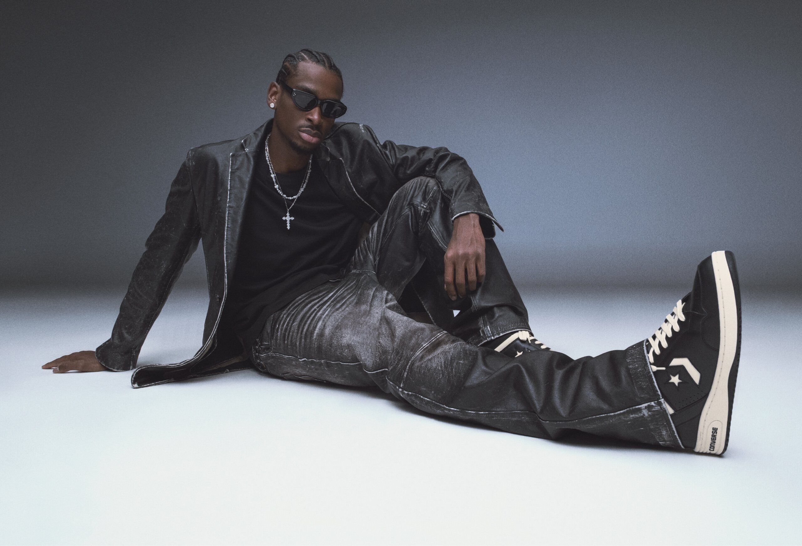 Shai Gilgeous-Alexander in leather jacket, sunglasses, jeans and Converse Weapon sneakers. He is sitting on white ground with one leg extended.