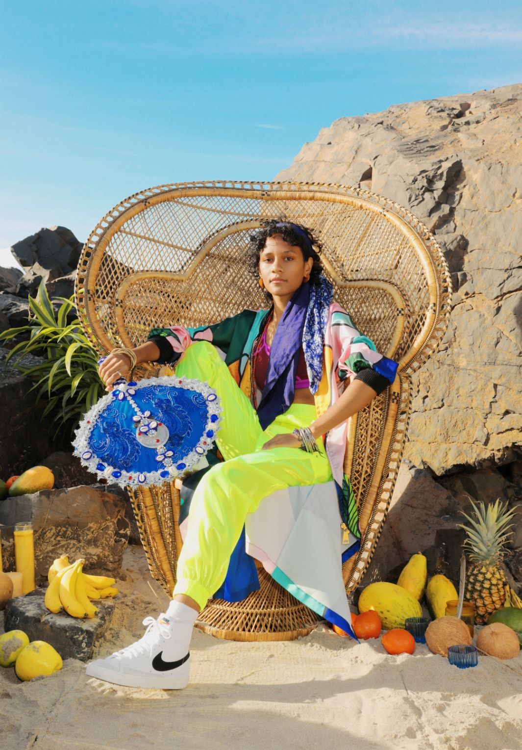 Djali Brown-Cepedain wicker chair on beach with colorful clothing. She is holding a decorated hand fan. There is fruit around the chair.