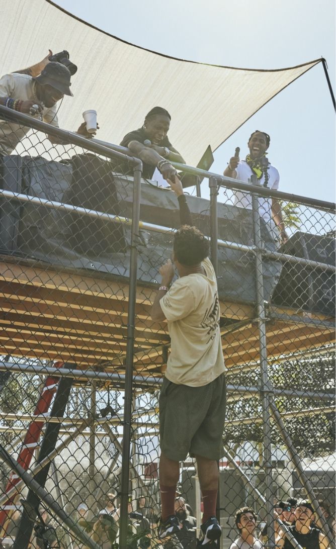 A person hands an item to someone on a raised platform behind a chain-link fence, while others on the platform watch and hold drinks.