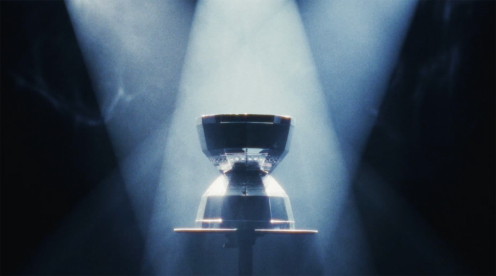 A metallic object, possibly a trophy or machine part, is displayed on a stand under four intersecting beams of spotlight in a dark, smoky setting.