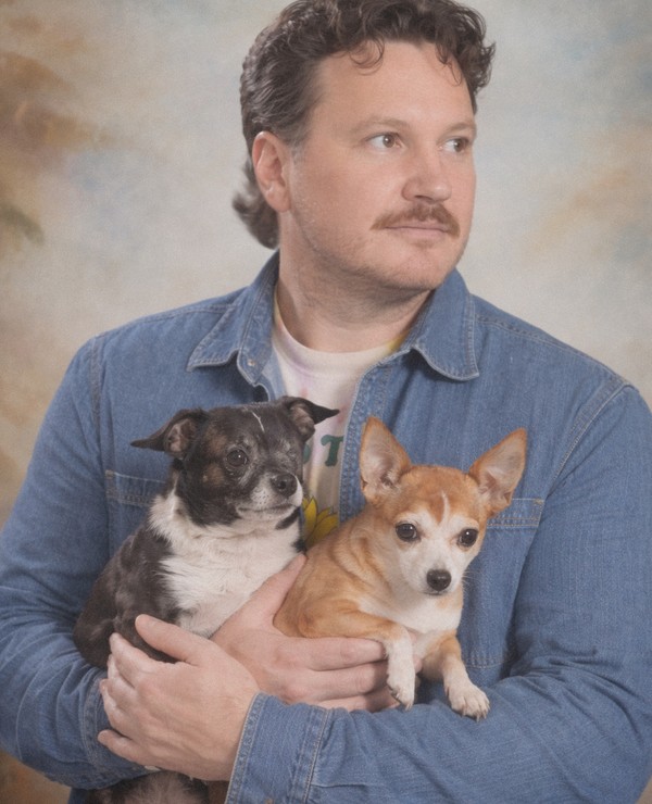 A man in a denim shirt holds two small dogs, one black and white and one tan and white, in front of a painted studio backdrop.