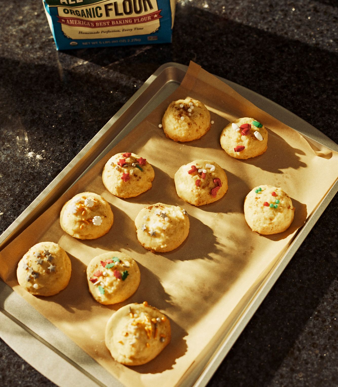 A baking tray lined with parchment paper holds ten round, decorated cookies; a bag of organic flour is visible in the background.
