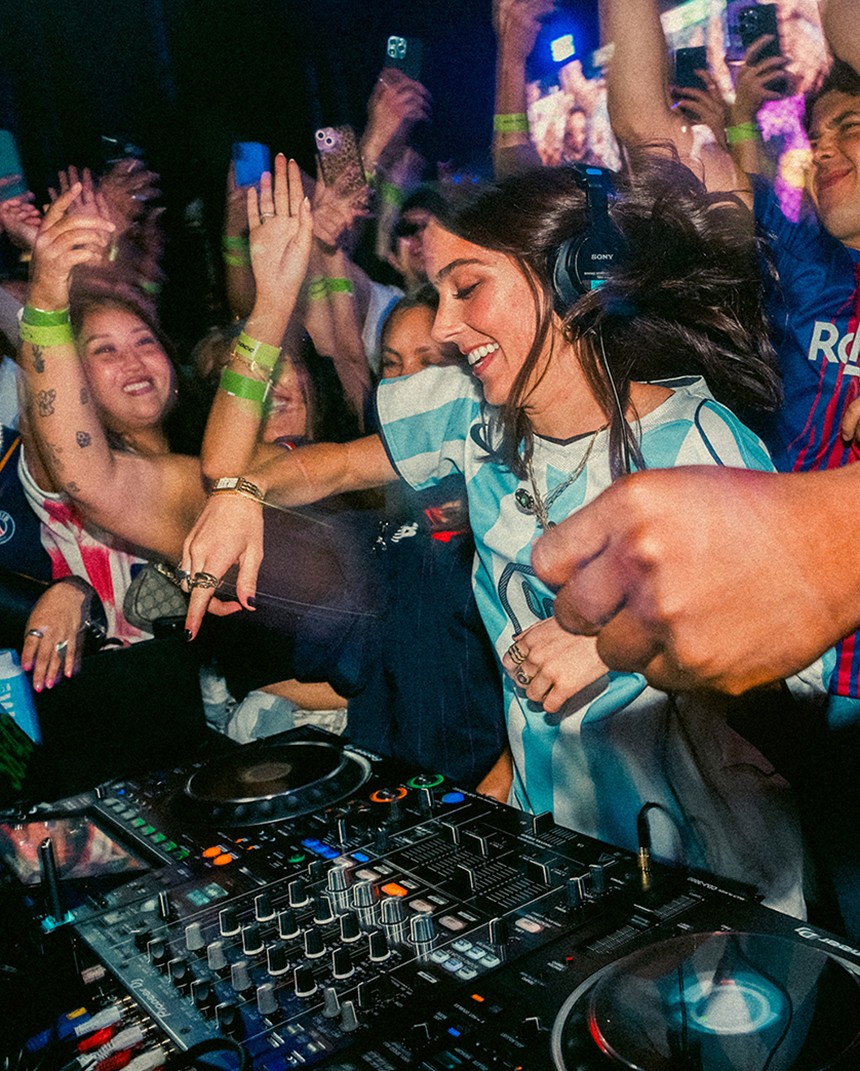 A DJ wearing headphones performs at a crowded event, smiling and mixing music as people around her dance and raise their hands in excitement.
