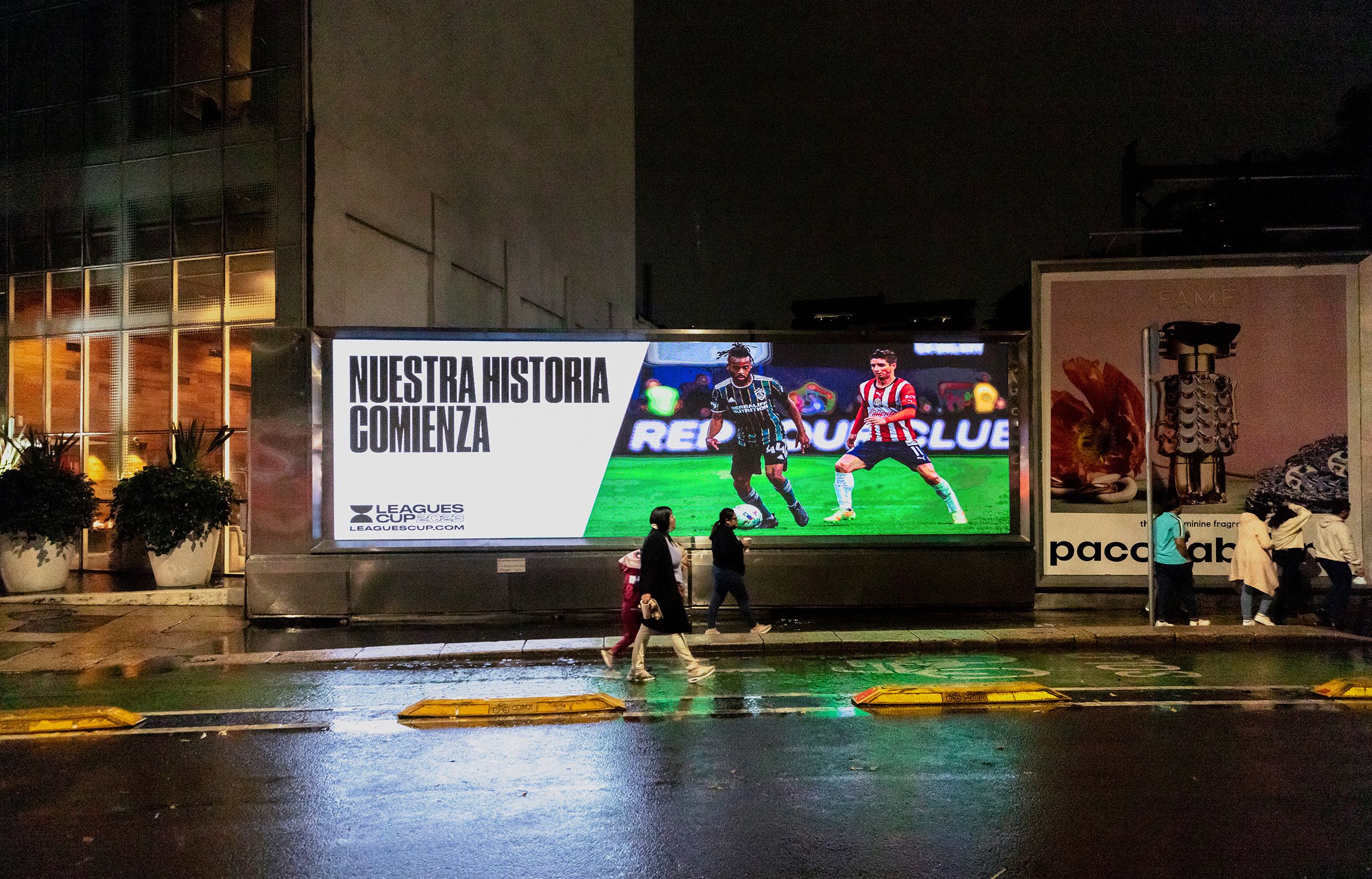 Large rectangular light up billboard along wet Mexico street at night. Two people walking in front of the billboard and a small gathering of people after the sign. Billboard reads Nestra Historia Comenza. Leagues Cup 2023.
